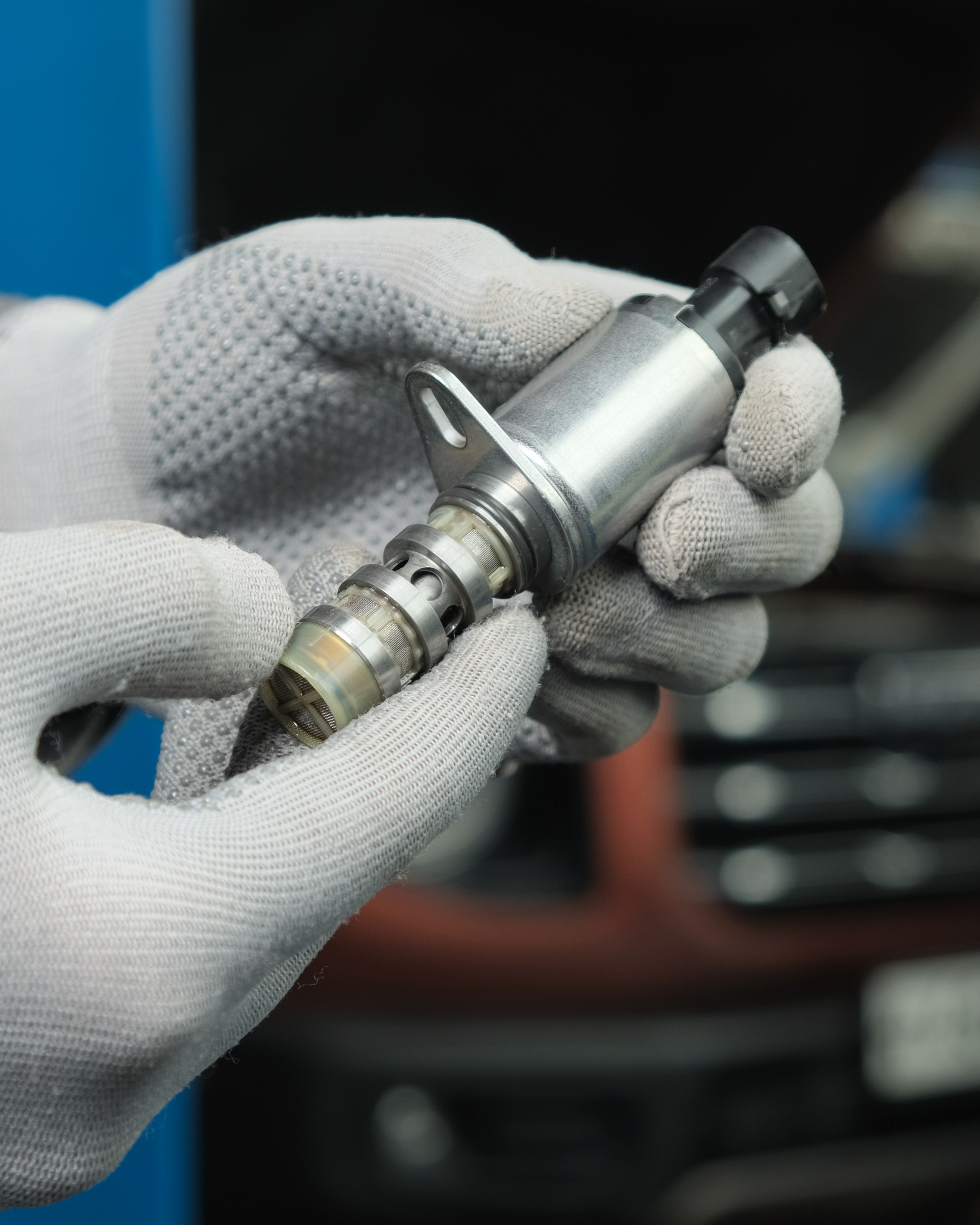Gloved hands holding a car part, a variable valve timing solenoid, with a worn tip, likely in a shop.
