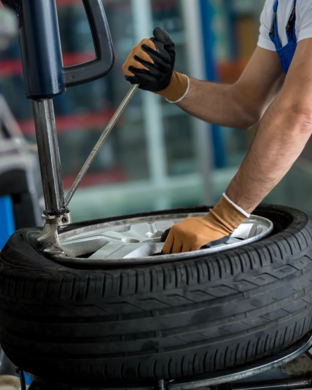 Mechanic using a tire machine to remove a tire from a wheel, wearing gloves, in a shop.