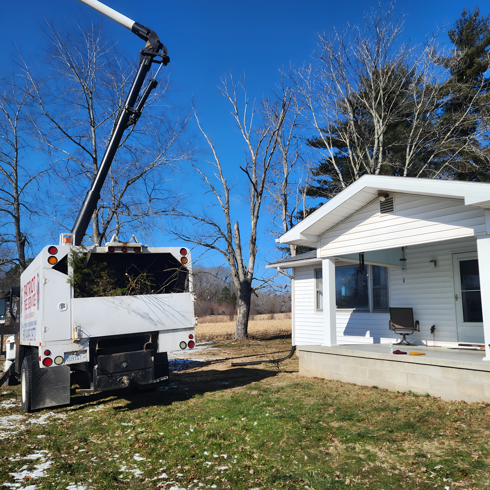 A tree service truck with a raised arm next to a white house, clearing debris on a sunny day.