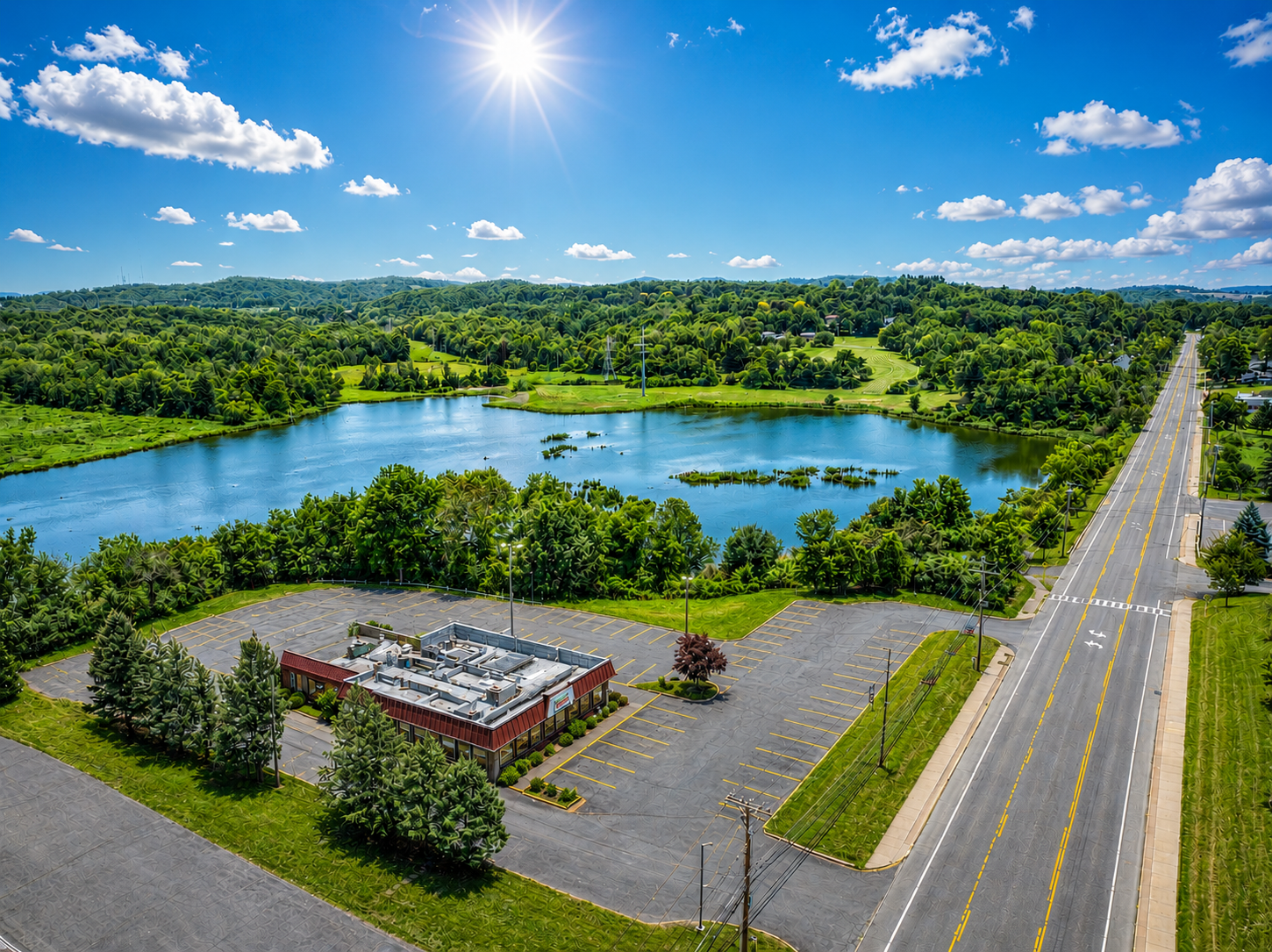 Aerial view of a lake, roadside, and small building surrounded by green trees under a bright blue sky.