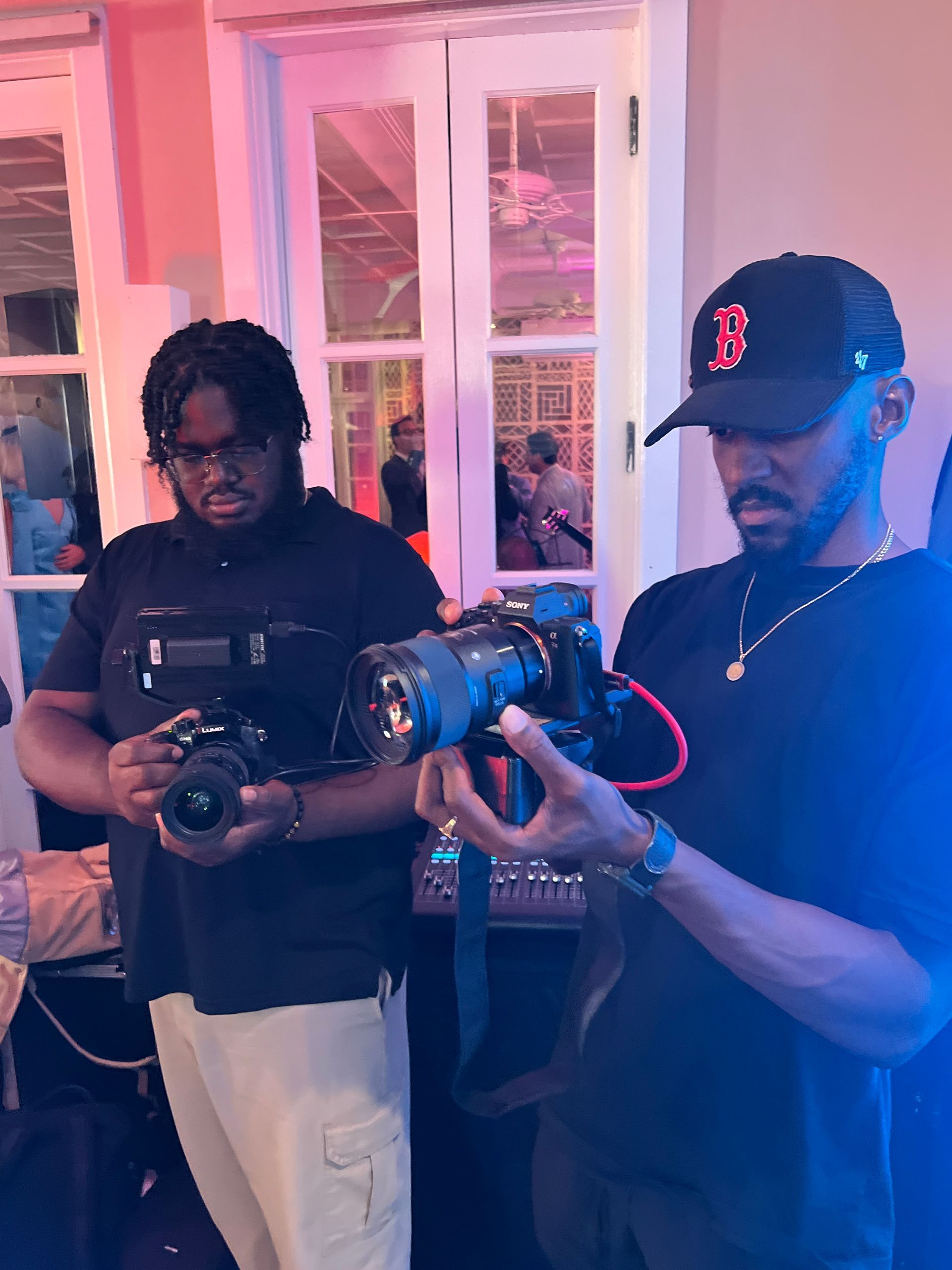 Two people holding camera rigs indoors, one adjusting a lens under pink lighting.