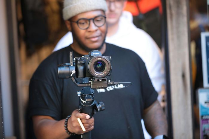 Person filming with a camera on a handheld gimbal indoors, wearing a beanie and glasses.
