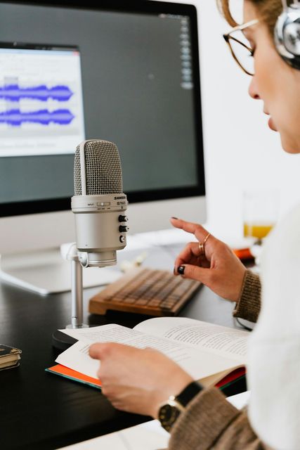 Person recording audio at a desk with a microphone, notebook, and computer showing sound waves.
