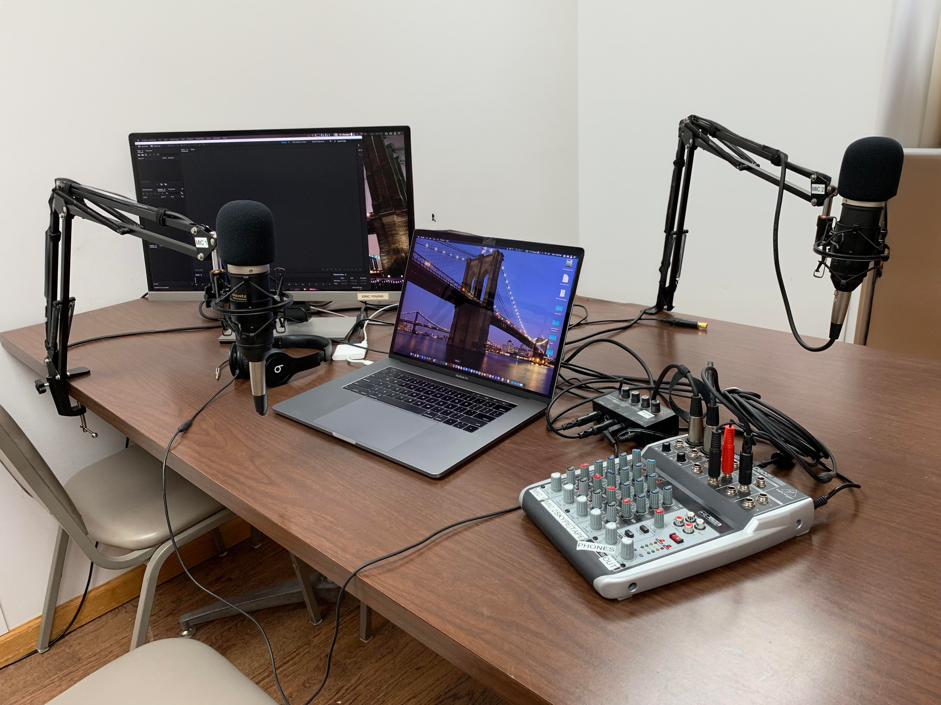 Podcast desk with laptop, monitors, microphones, and audio mixer on a wooden table.