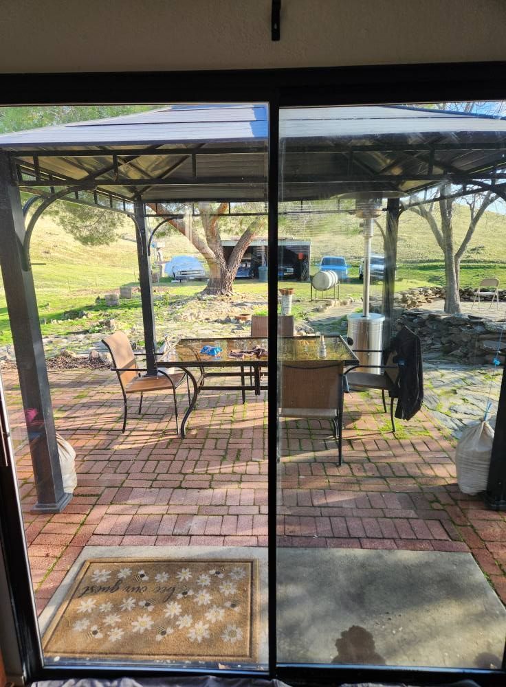 View through a sliding glass door to a patio with chairs, brick pavers, and a pergola in a grassy yard.