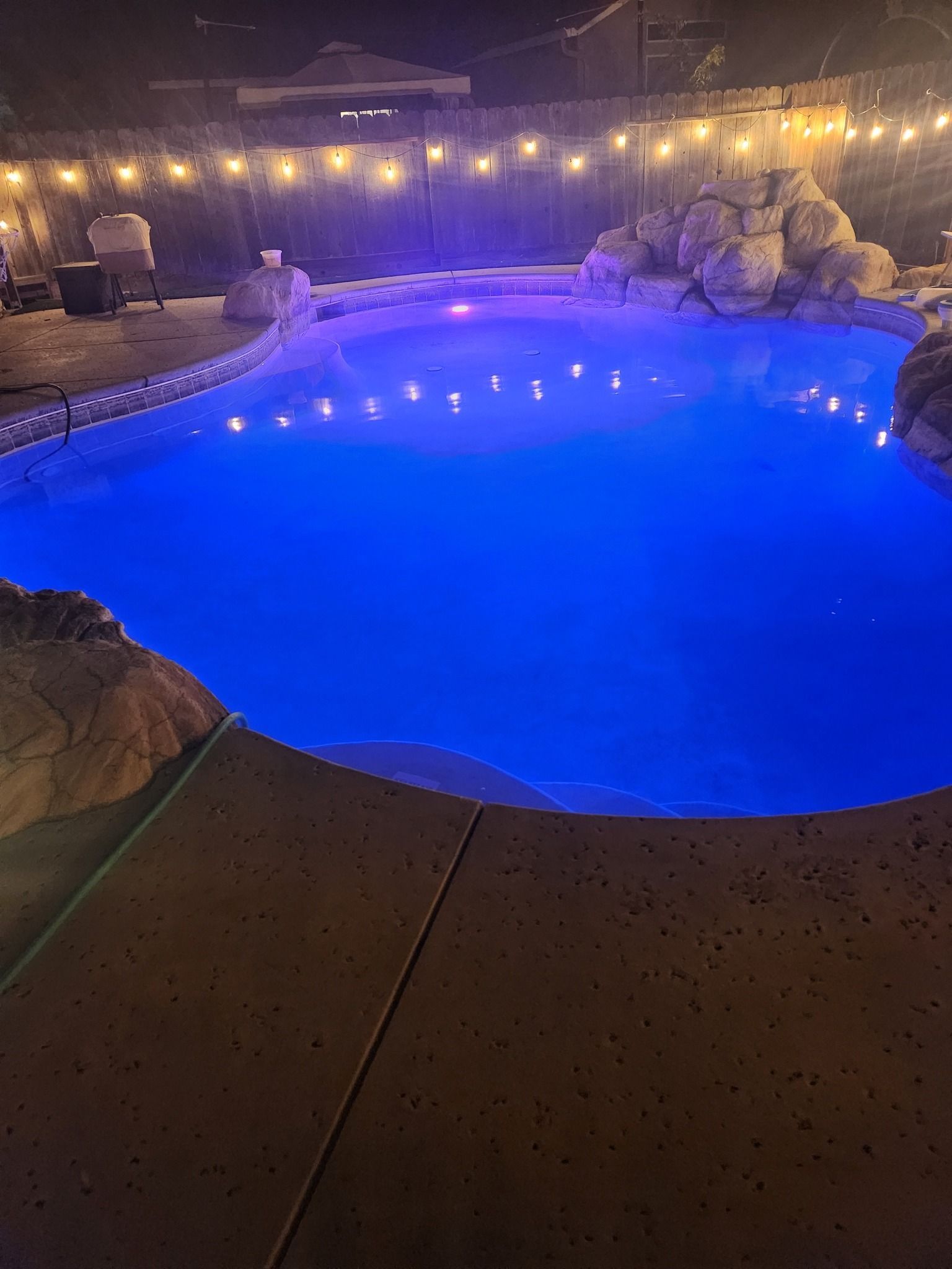 Blue-lit backyard pool at night with string lights and surrounding rocks