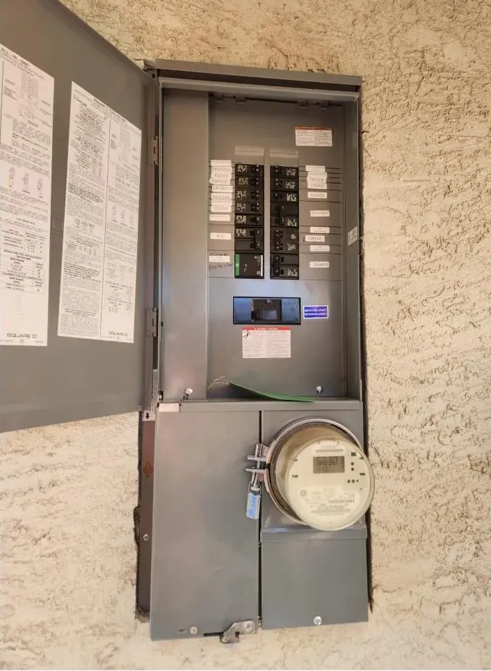 Electrical breaker panel with utility meter on a wall, open cover showing labels and switches