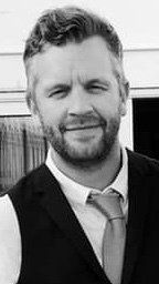 A man with a beard is wearing a vest and tie and smiling in a black and white photo.