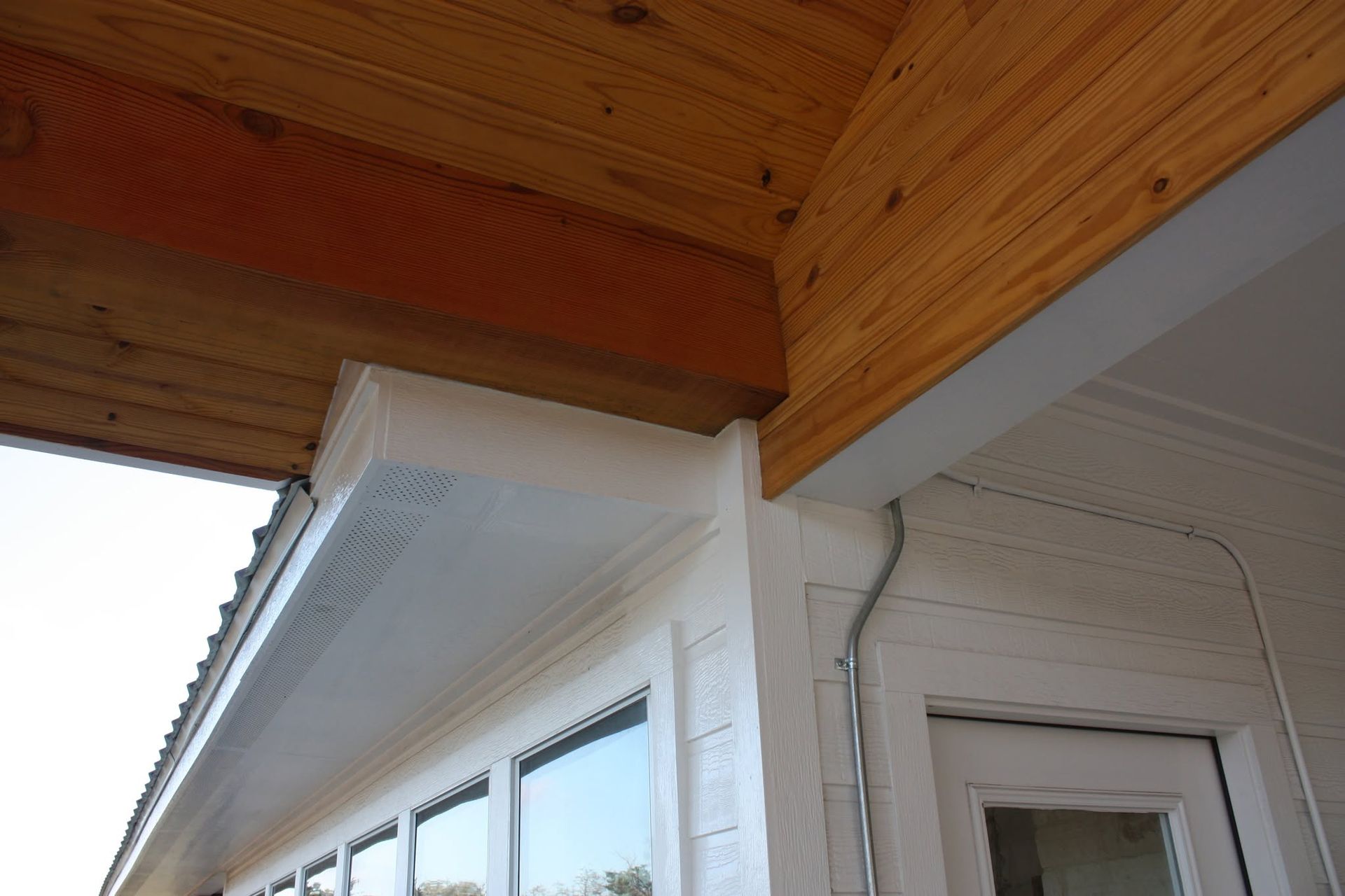 Wood-paneled porch ceiling over white house siding and windows, with a corner gutter and doorway detail