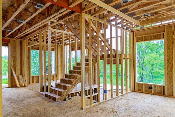 Unfinished wooden house interior with exposed framing, stairs, and large window openings overlooking greenery