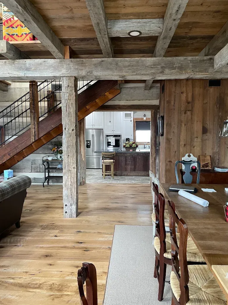 Rustic open-plan living room with exposed beams, wooden staircase, and kitchen in the background