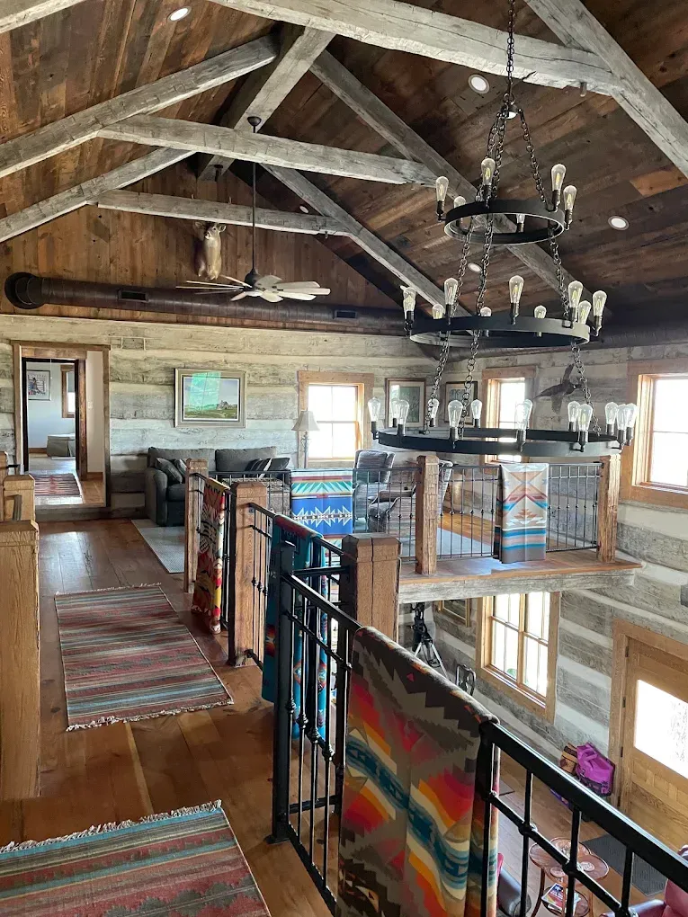 Rustic loft interior with exposed beams, stone fireplace, black railing, and colorful woven blanket.