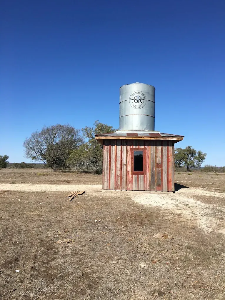 Small weathered red shed with a metal water tank on top in a dry rural field under a clear blue sky