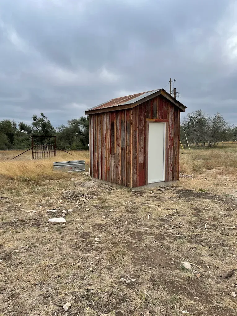 Small weathered wooden shed with a white door in a dry, open field under a cloudy sky