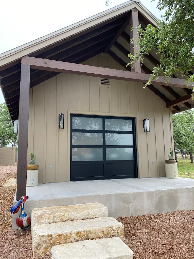 Small beige garage with black door, timber roof beams, and concrete steps in a landscaped yard.