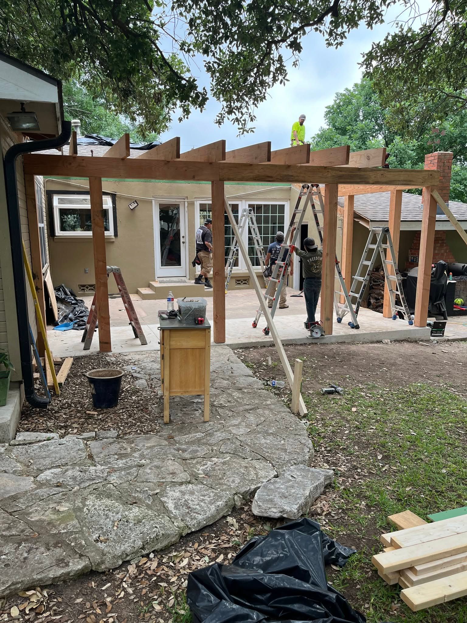 Wooden pergola under construction in a yard, with tools, lumber, and a house in the background.