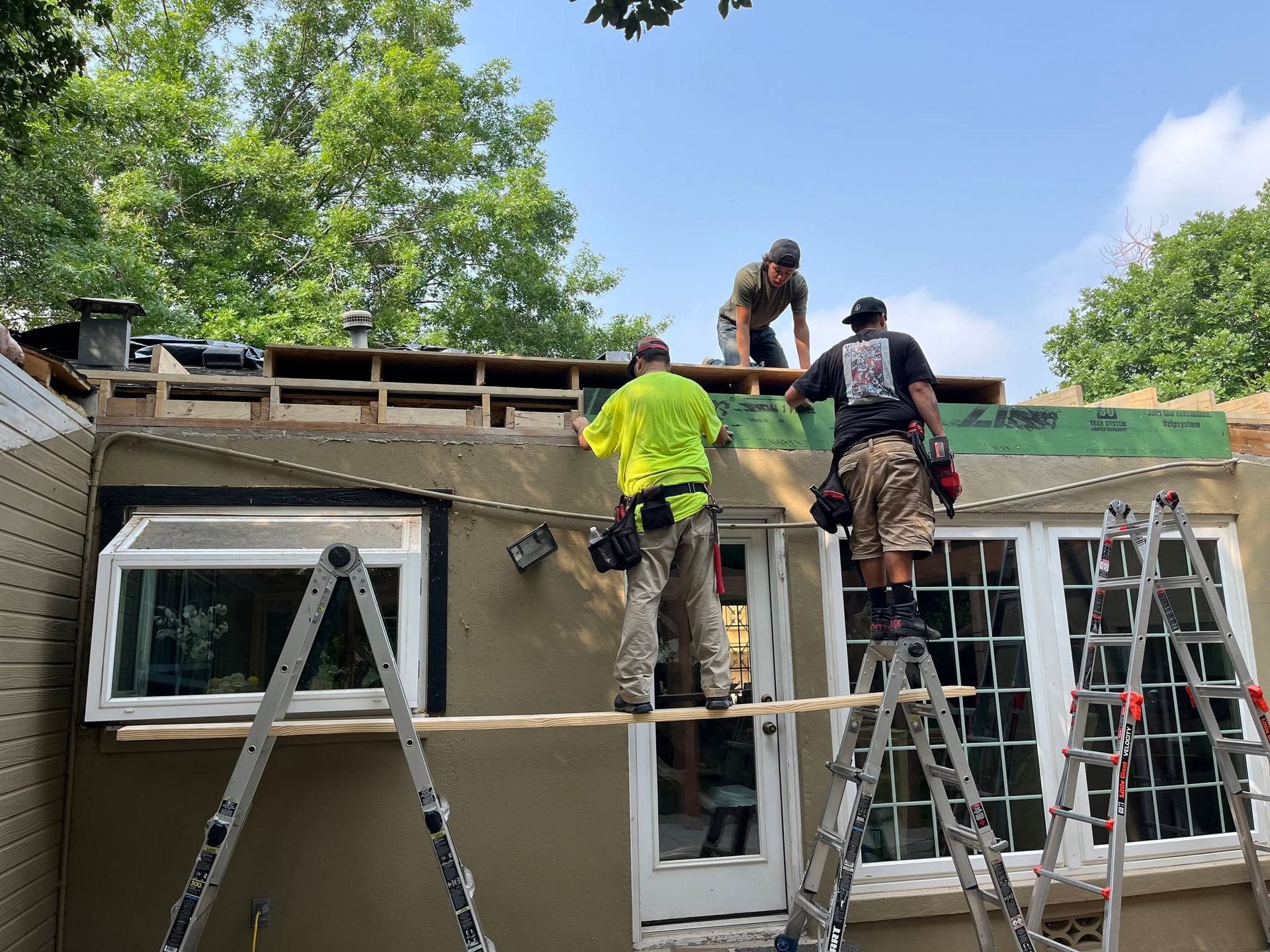 Three workers on ladders repairing a house roof and exterior walls under a blue sky.