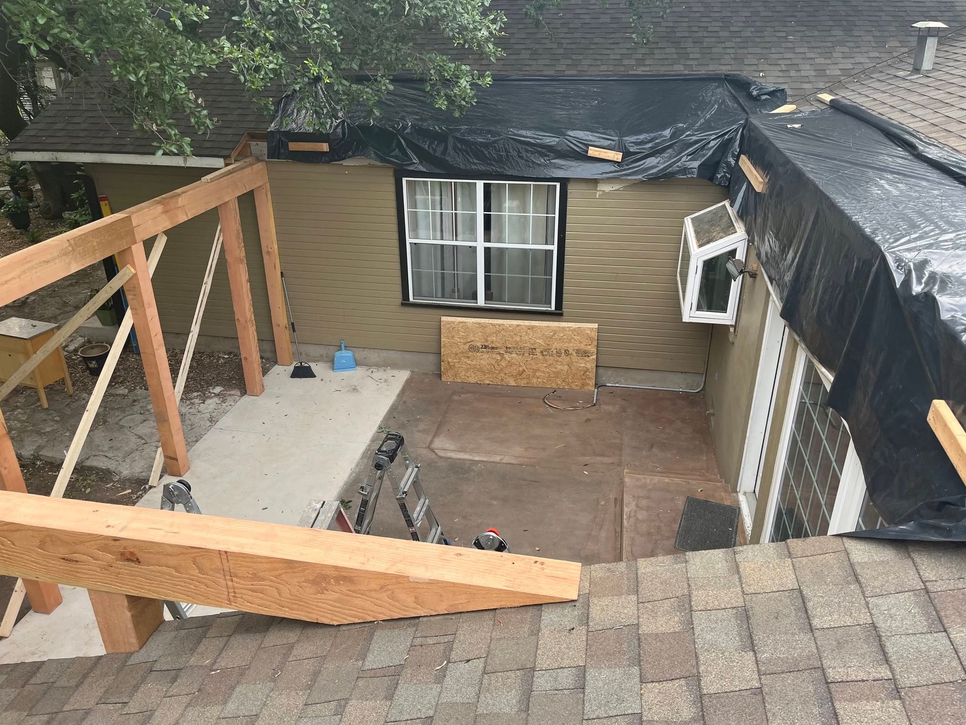 Roofing work overlooking a backyard patio with ladders, plywood, and tarps on the roof