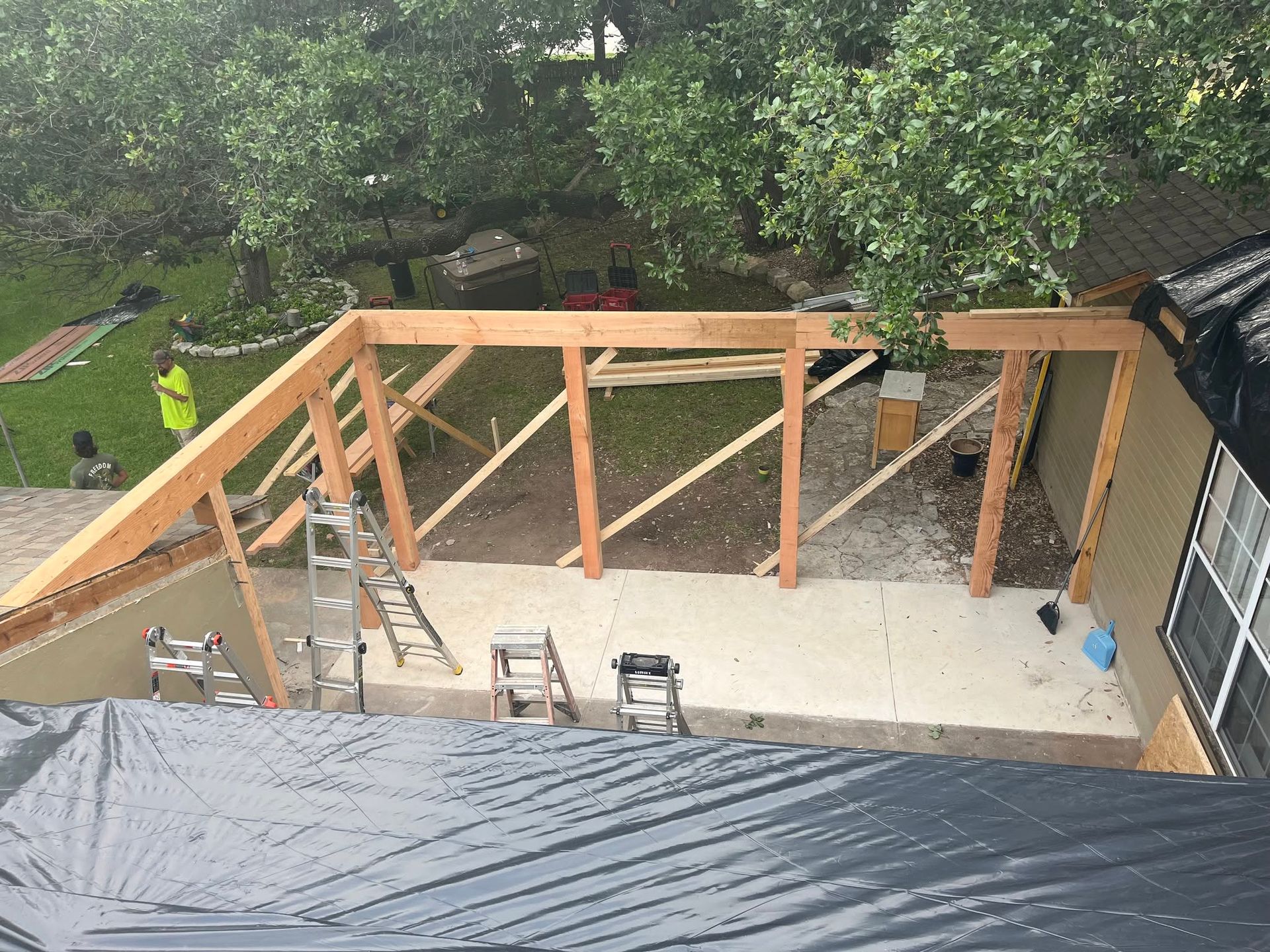 Wooden roof framing under construction beside a house, with ladders and a tarp-covered area below.