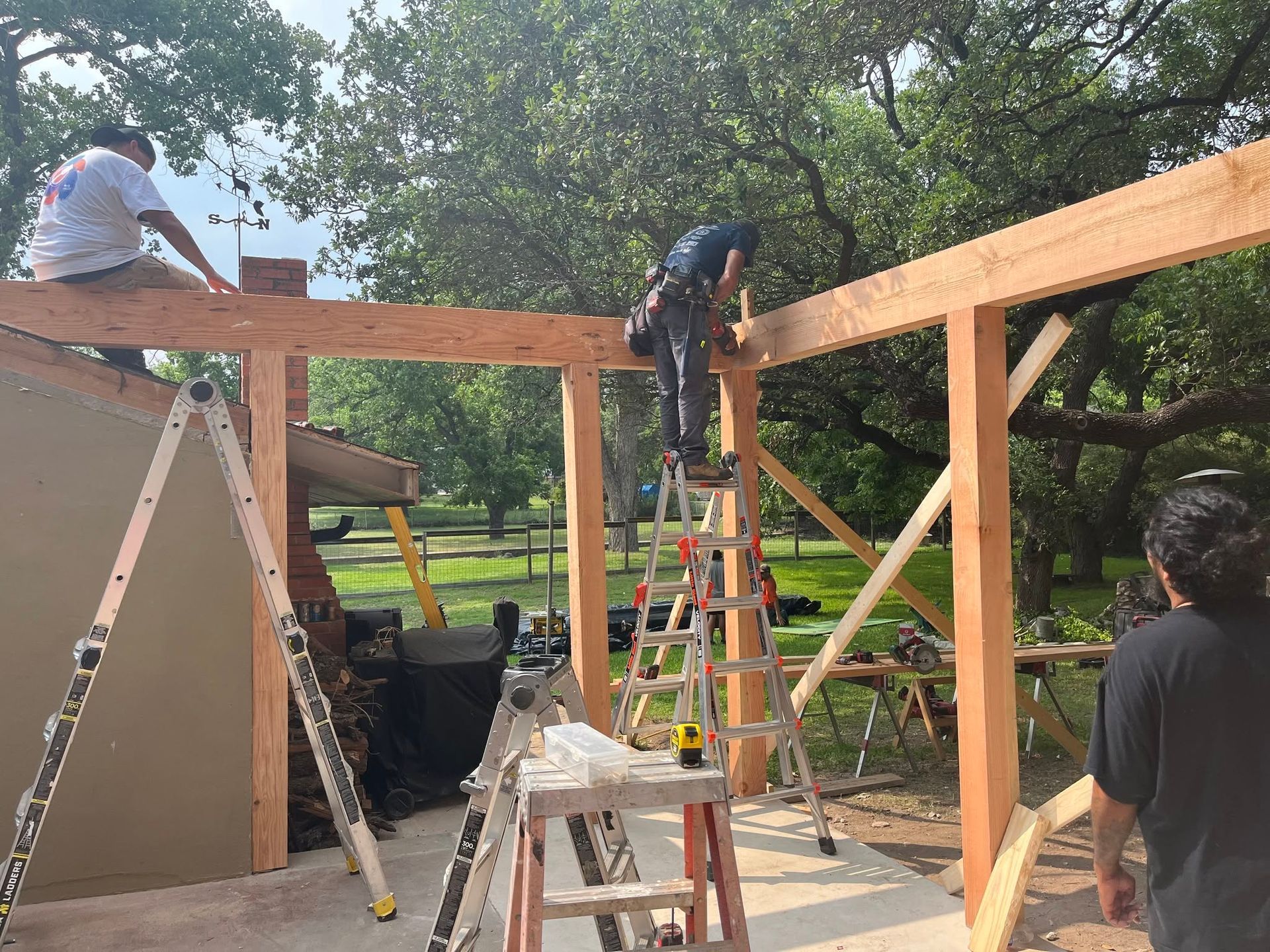 Workers installing wooden beams on a structure frame outdoors, with ladders and trees in the background