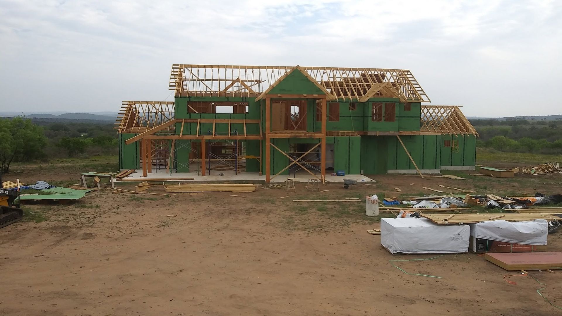 Partially built green house with exposed wooden roof framing on a dirt lot under cloudy sky