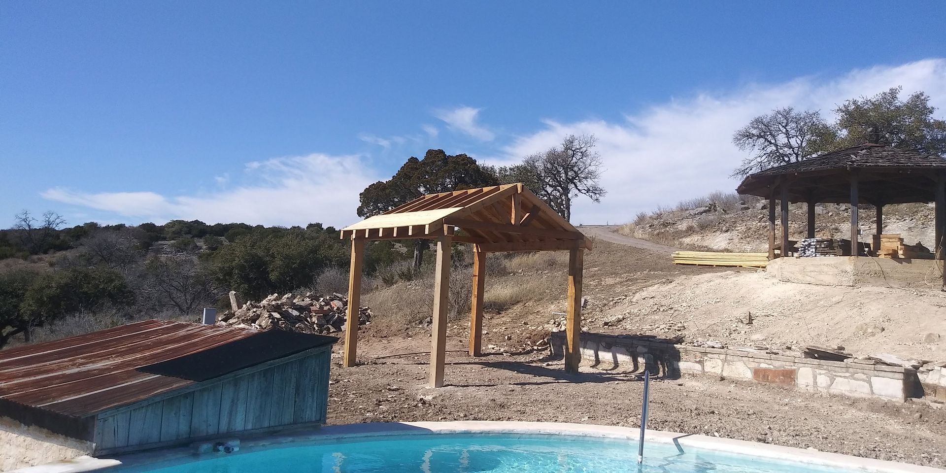 Outdoor rustic structure with wooden posts, stone ground, red-roofed shed, and dry hillside under a blue sky