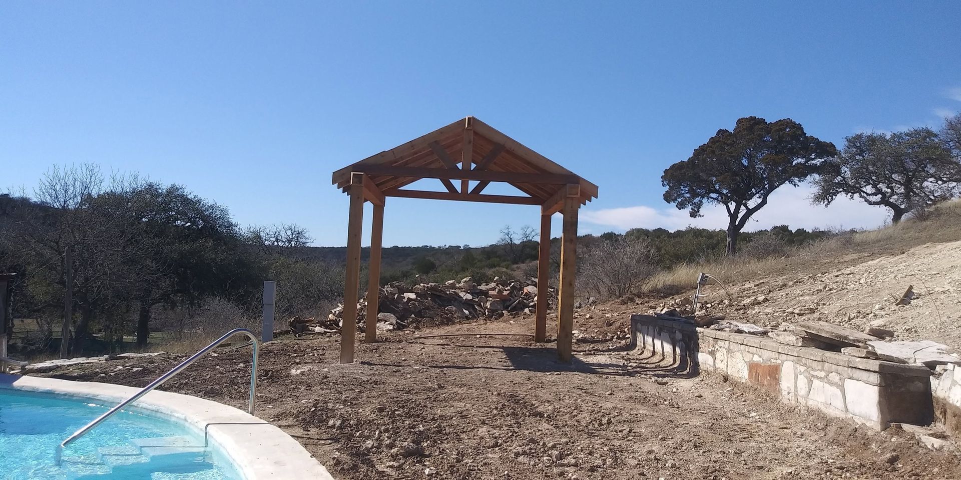 Wooden pergola beside a blue pool in a dry hillside landscape with scattered trees