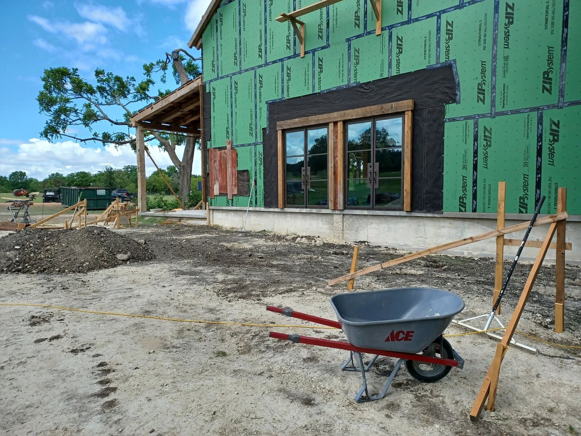 Partially built house with green sheathing, a wheelbarrow, and tools on a gravel construction site