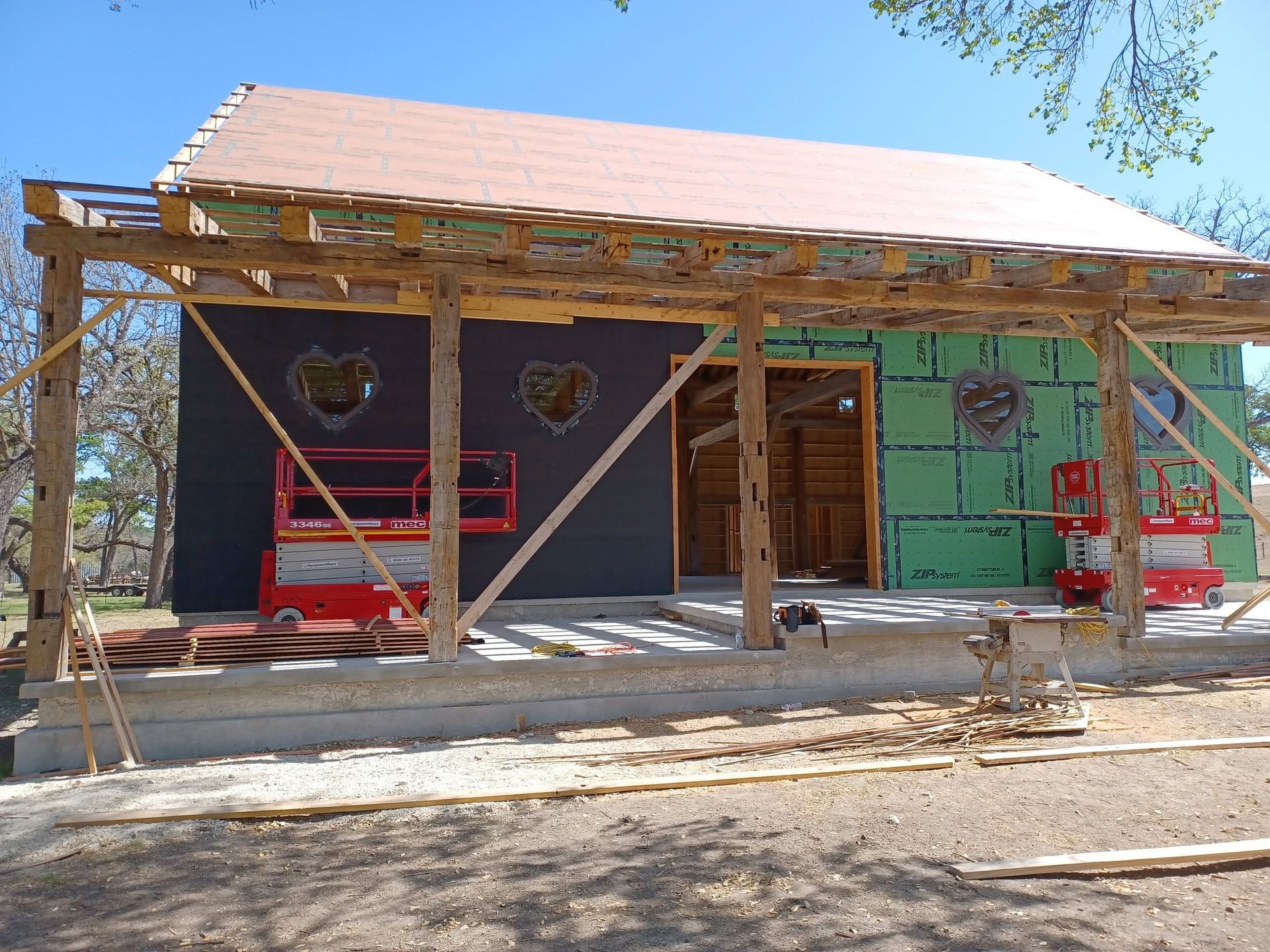 Partially built house with exposed framing, green and dark siding, and red scaffolding on a sunny day