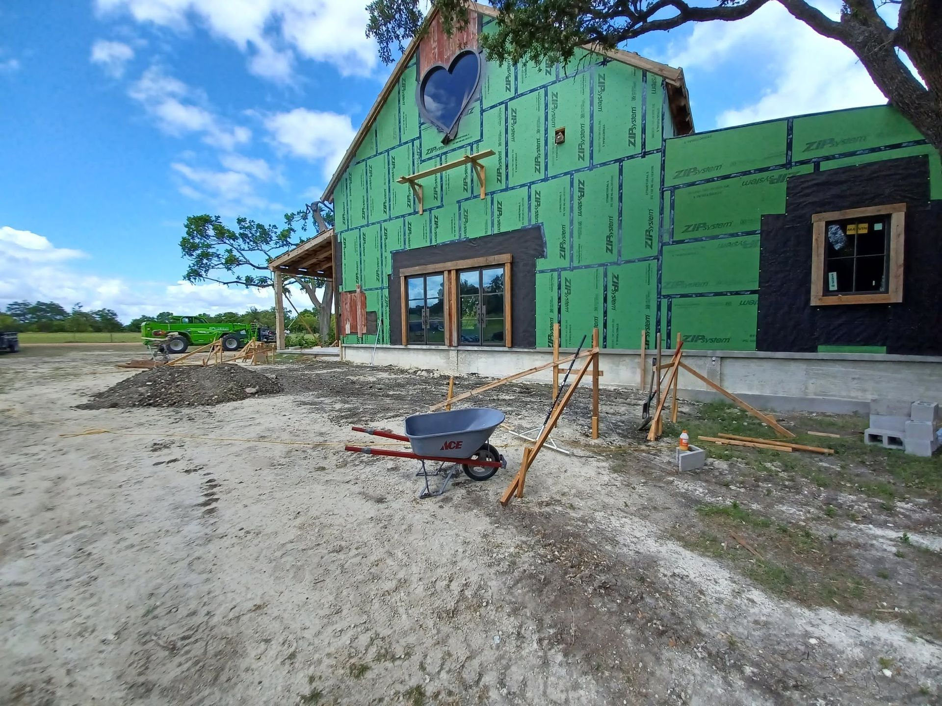 Green house under construction with exposed framing, sandy yard, and a wheelbarrow in front