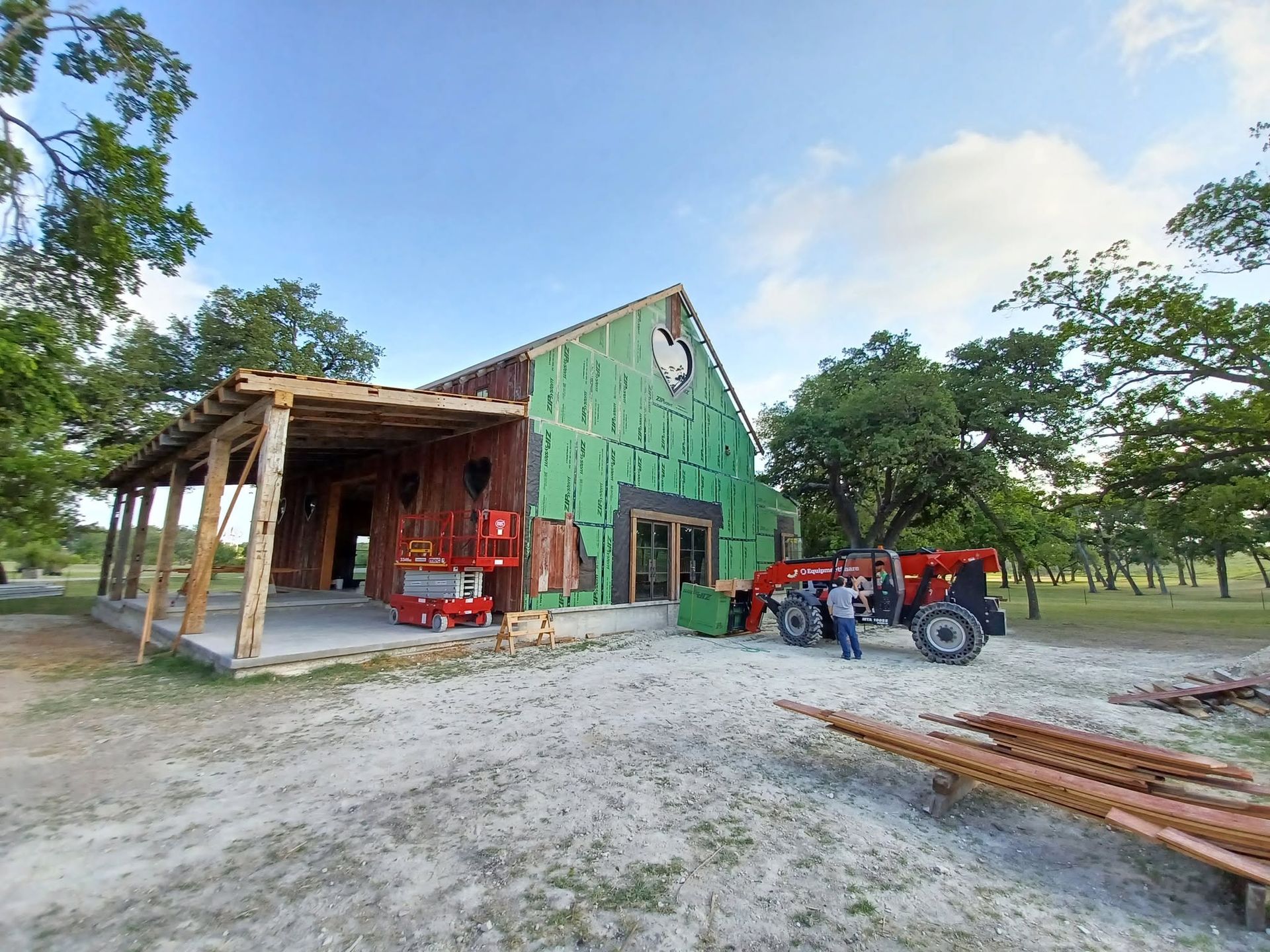 Green barn with a porch and a red tractor parked outside on a rural dirt lot, surrounded by trees.