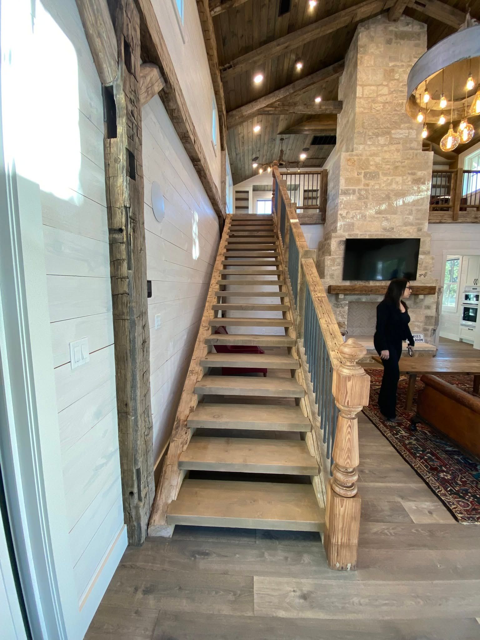 Tall wooden staircase in a rustic lodge interior with stone fireplace and chandelier.