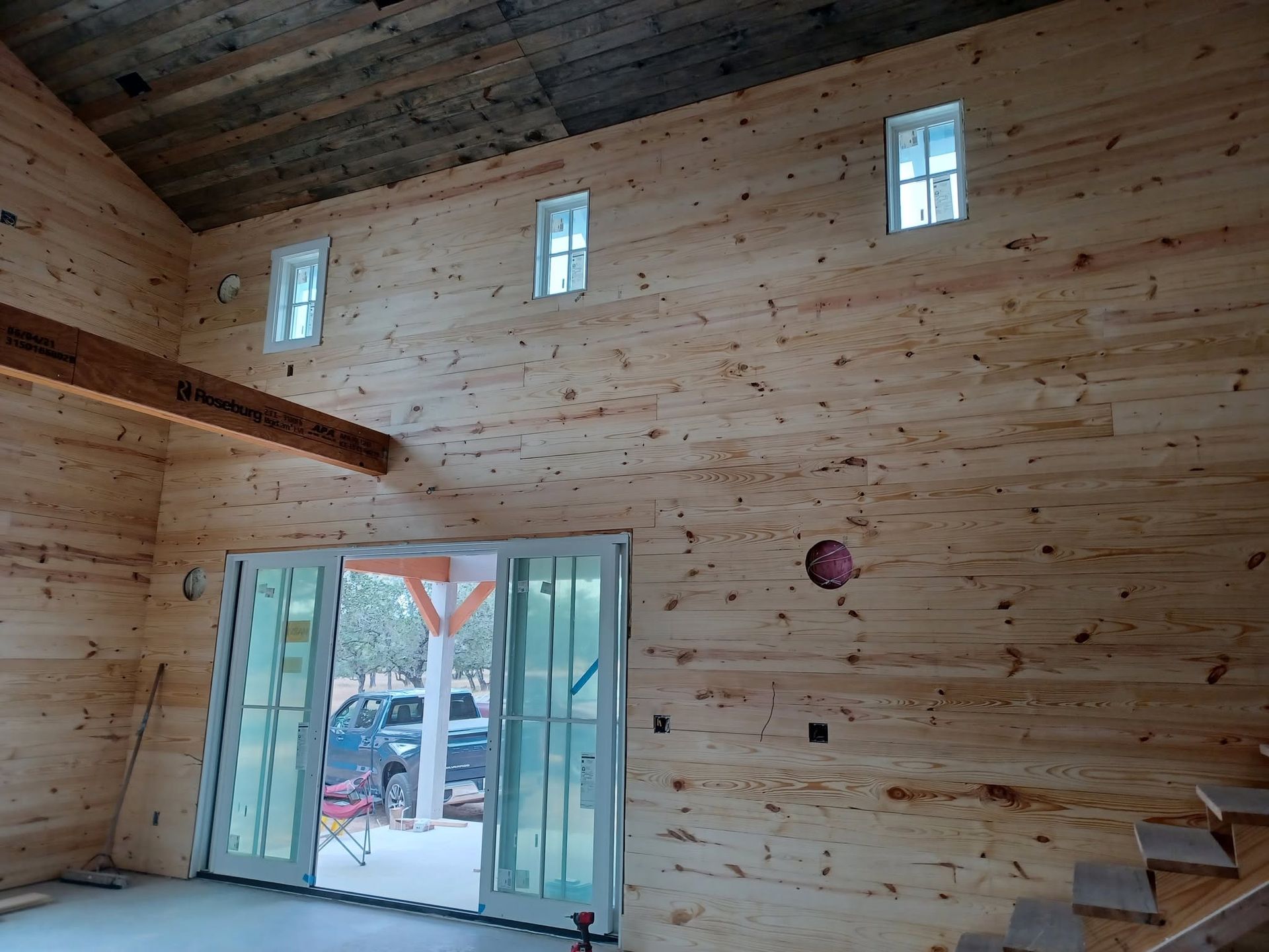 Spacious unfinished wood interior with tall windows, open French doors, and a staircase at right.