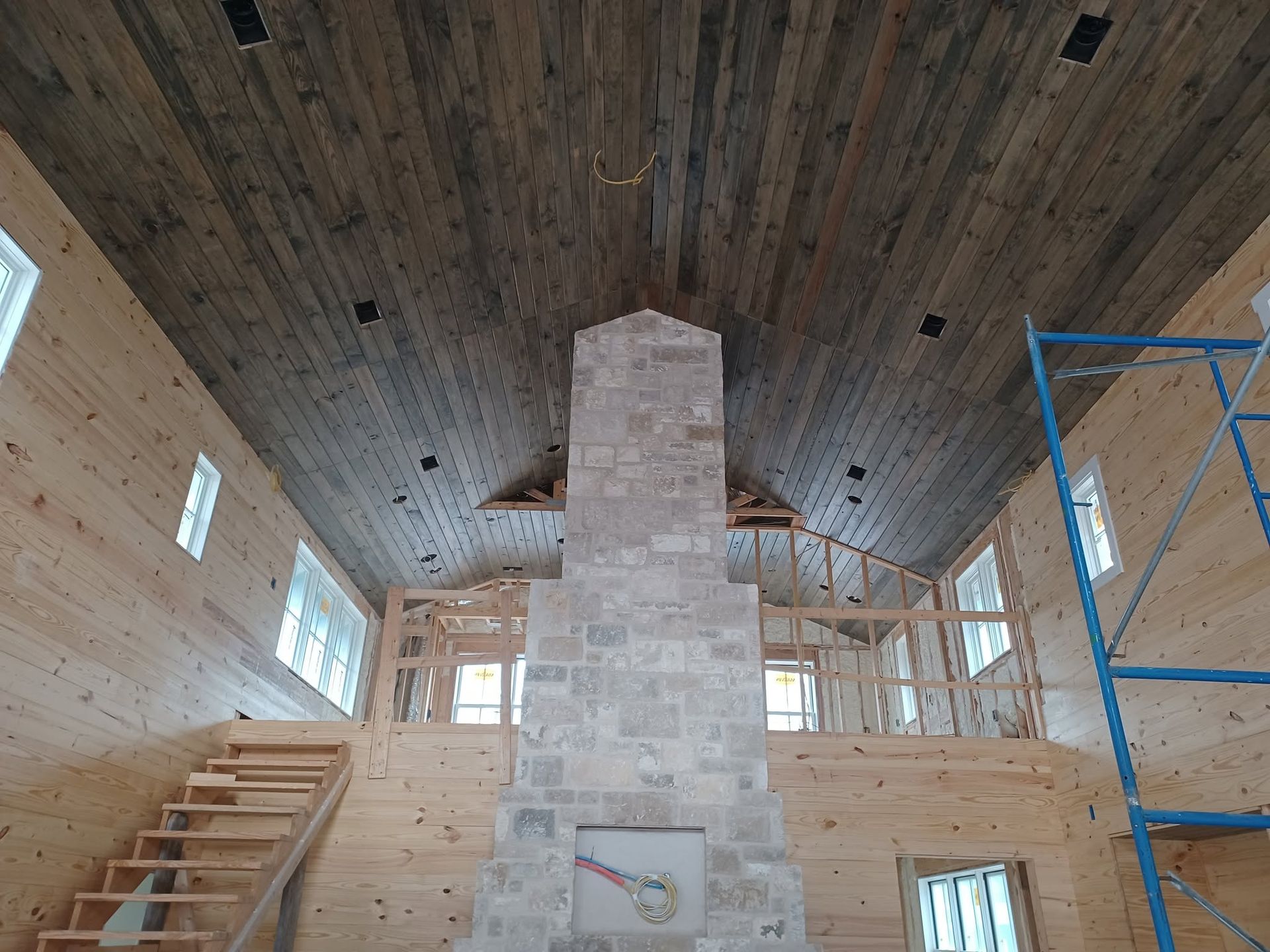 Unfinished interior with high wood ceiling, central stone fireplace, stairs, windows, and blue scaffolding.