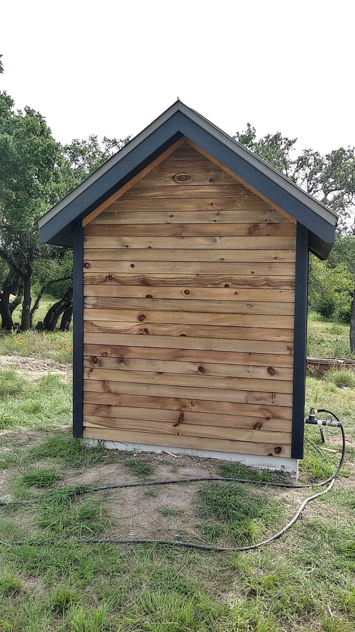 Small wooden shed with a dark roof in a grassy outdoor area, viewed from the front.