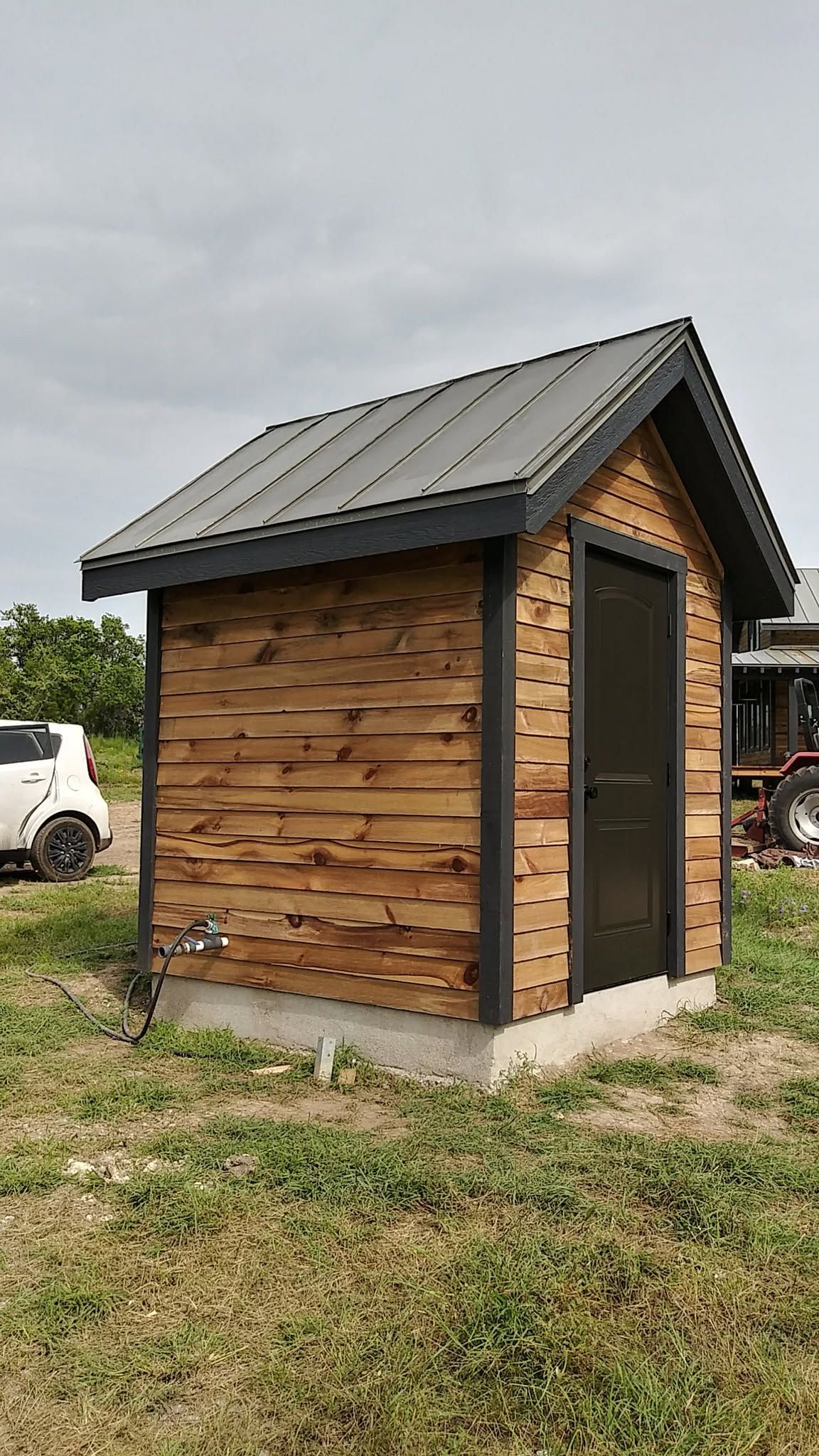 Small wooden shed with a dark metal roof on a grassy lot, with a white vehicle in the background