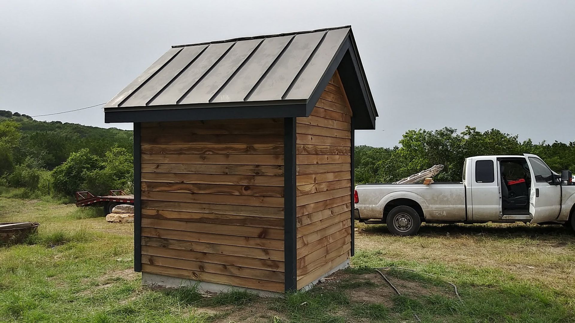 Small wooden shed with metal roof in a grassy field, with a white pickup truck parked behind it.