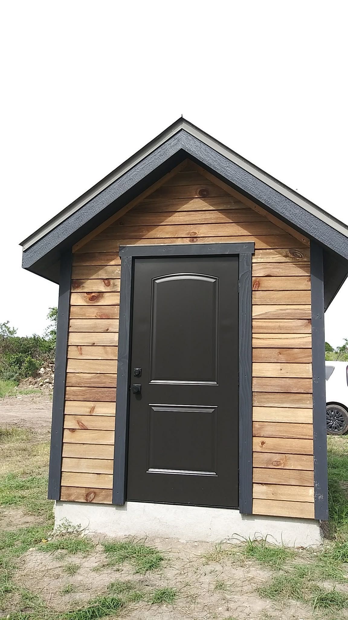 Small wooden shed with a black door and gray roof on a grassy outdoor lot.