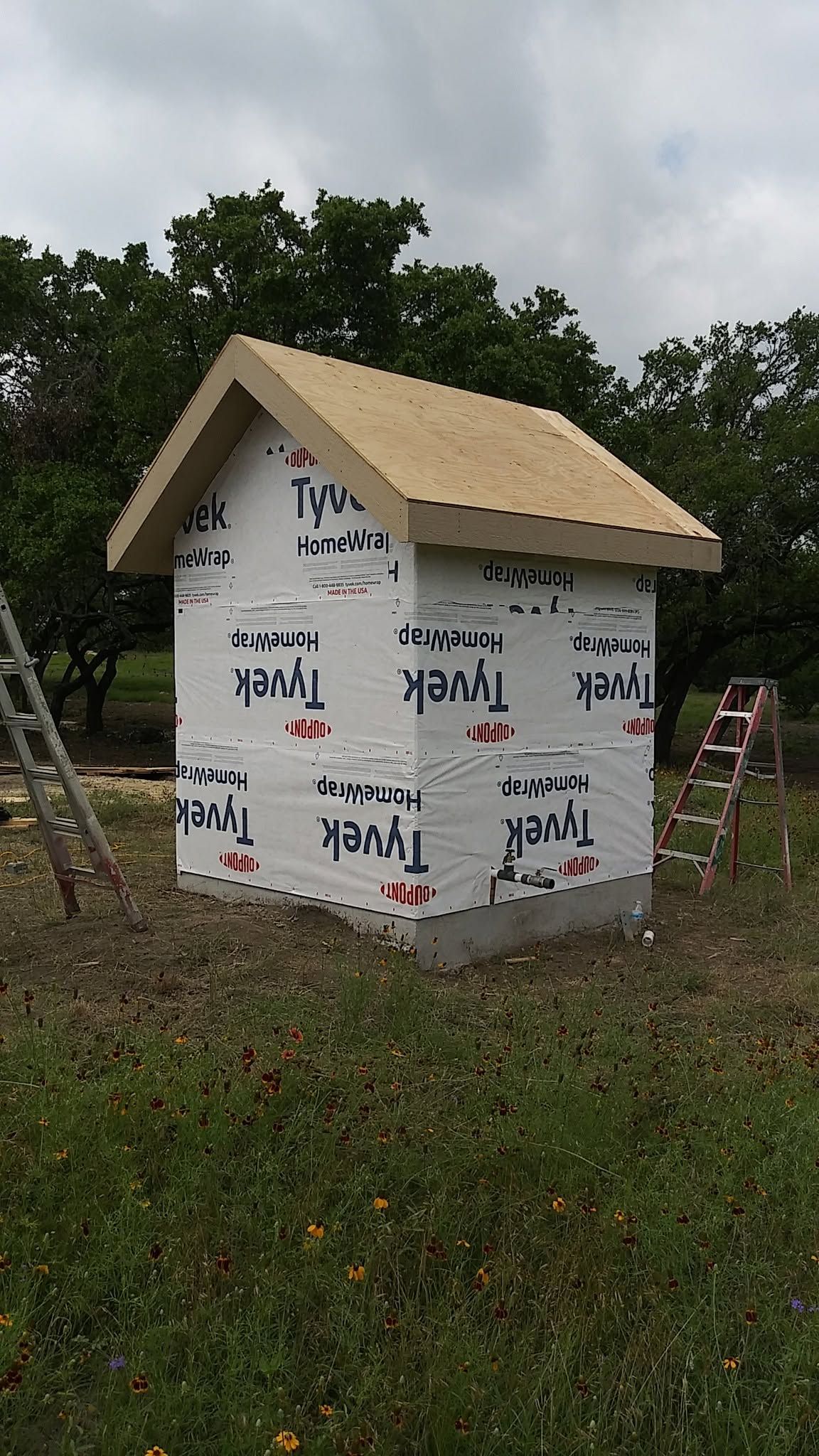 Small white shed wrapped in protective sheeting, with a tan roof, standing in a grassy outdoor area.