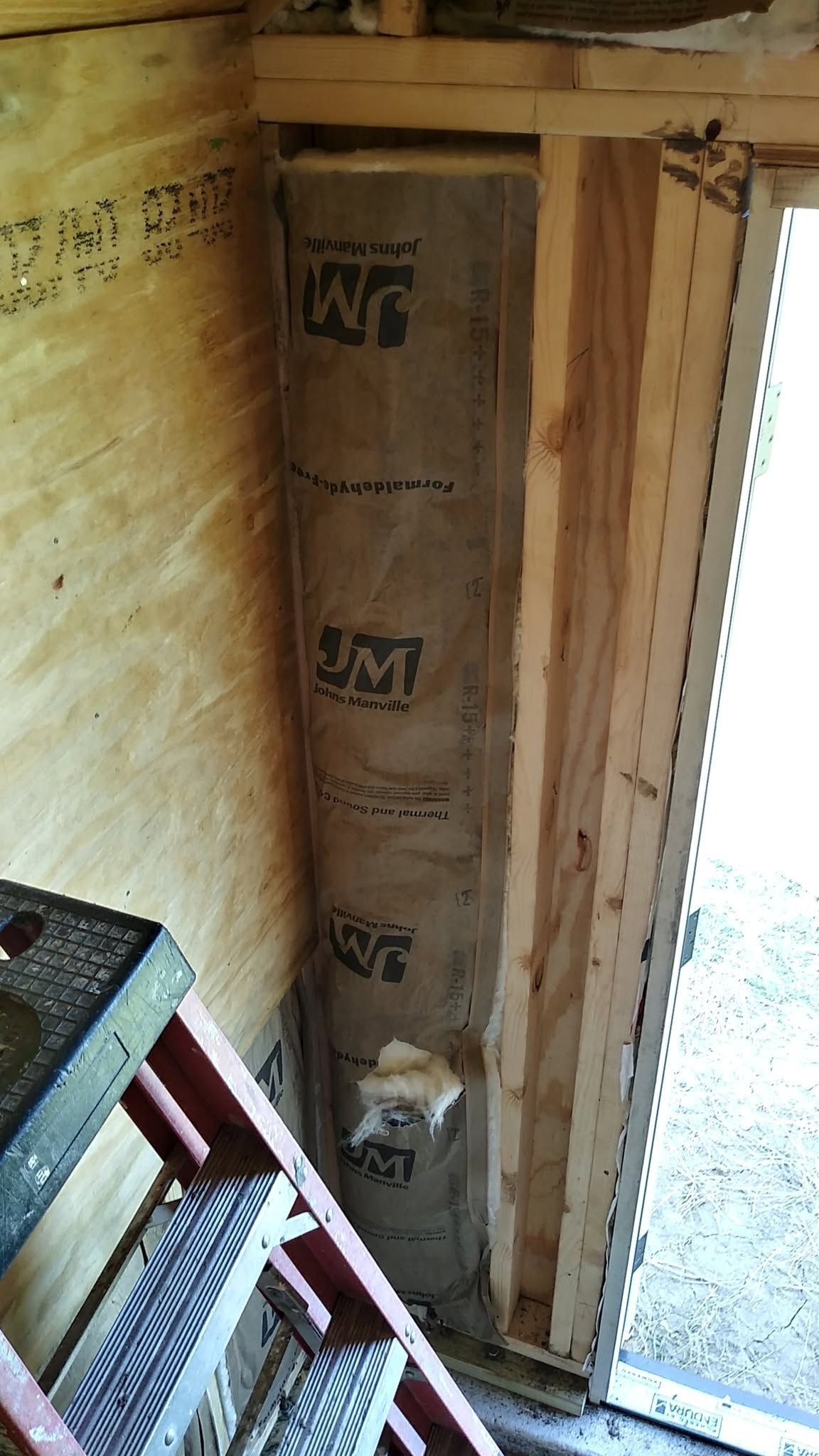 Insulated wooden wall cavity with exposed framing beside a doorway and ladder indoors