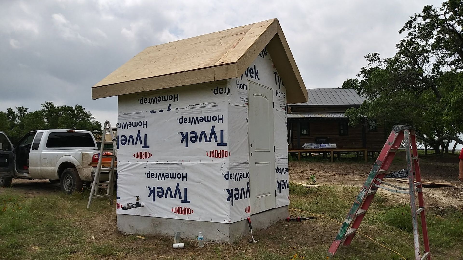 Small white shed under construction with wrapped siding, plywood roof, ladder, and truck in a grassy yard.