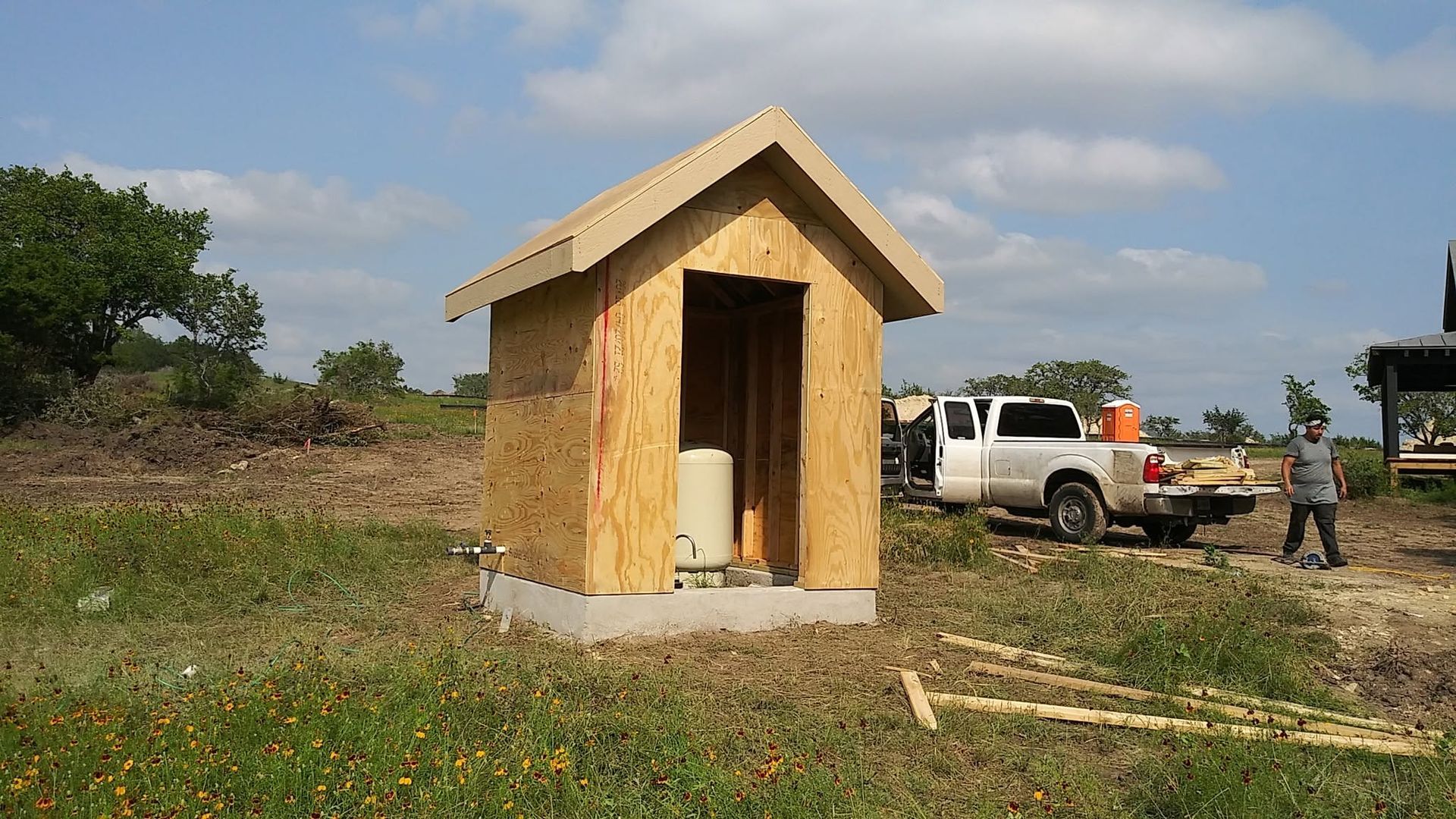 Small plywood shed in a grassy field with a white pickup truck and a person in the background.