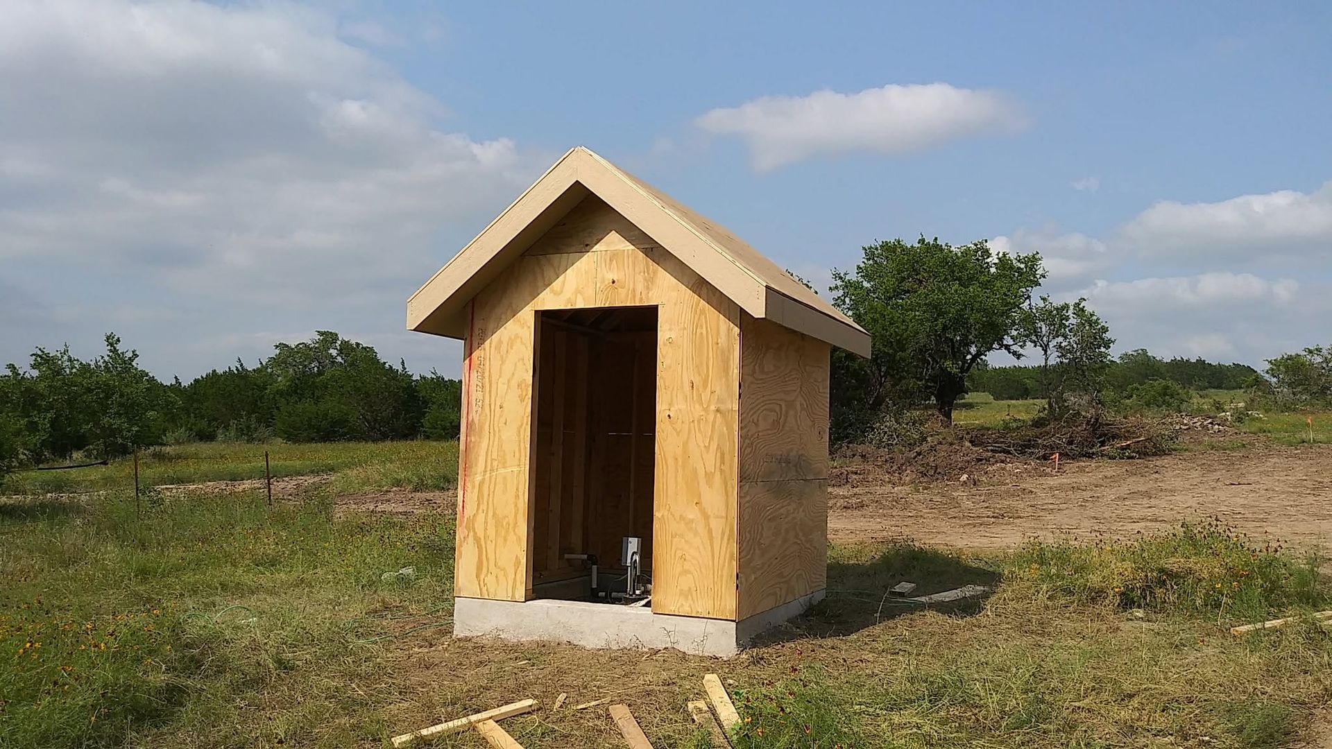 Small tan shed with a peaked roof in a grassy field under a partly cloudy sky.