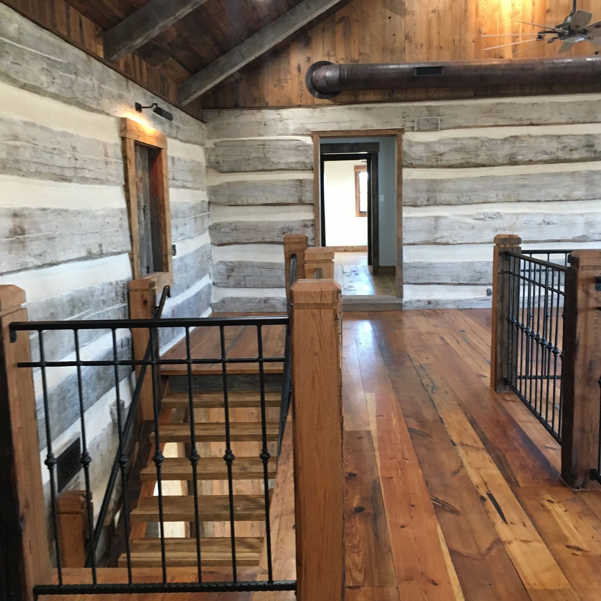 Rustic log cabin interior with wood floors, black railings, and an open doorway to another room.
