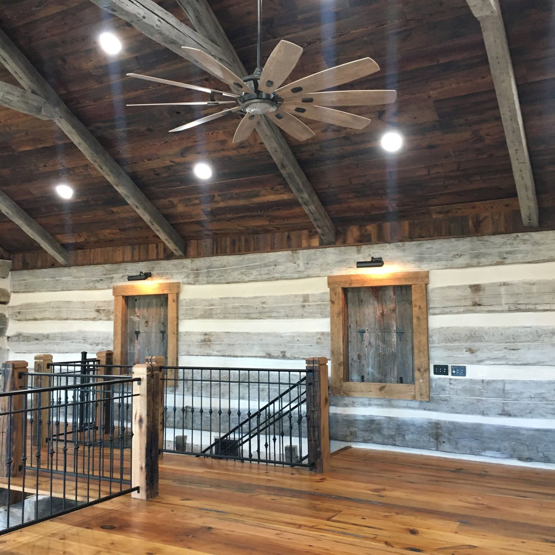 Rustic loft interior with wood beams, ceiling fan, railings, and large wooden doors above a hardwood floor