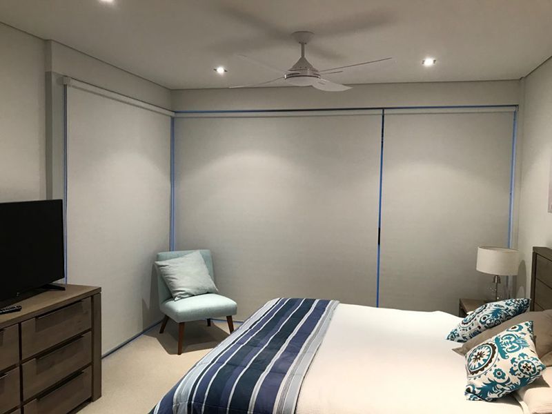 Bedroom With White Roller Shades, Furniture, Ceiling Fan, And Bed — Winning Blinds in Cardiff, NSW