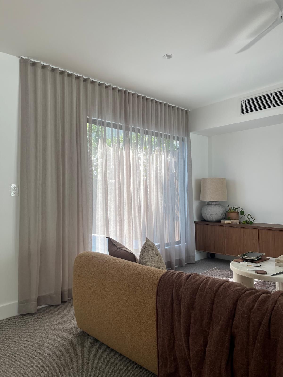 Modern living room with a tan sofa, brown throw, large curtained window, and sideboard decor.