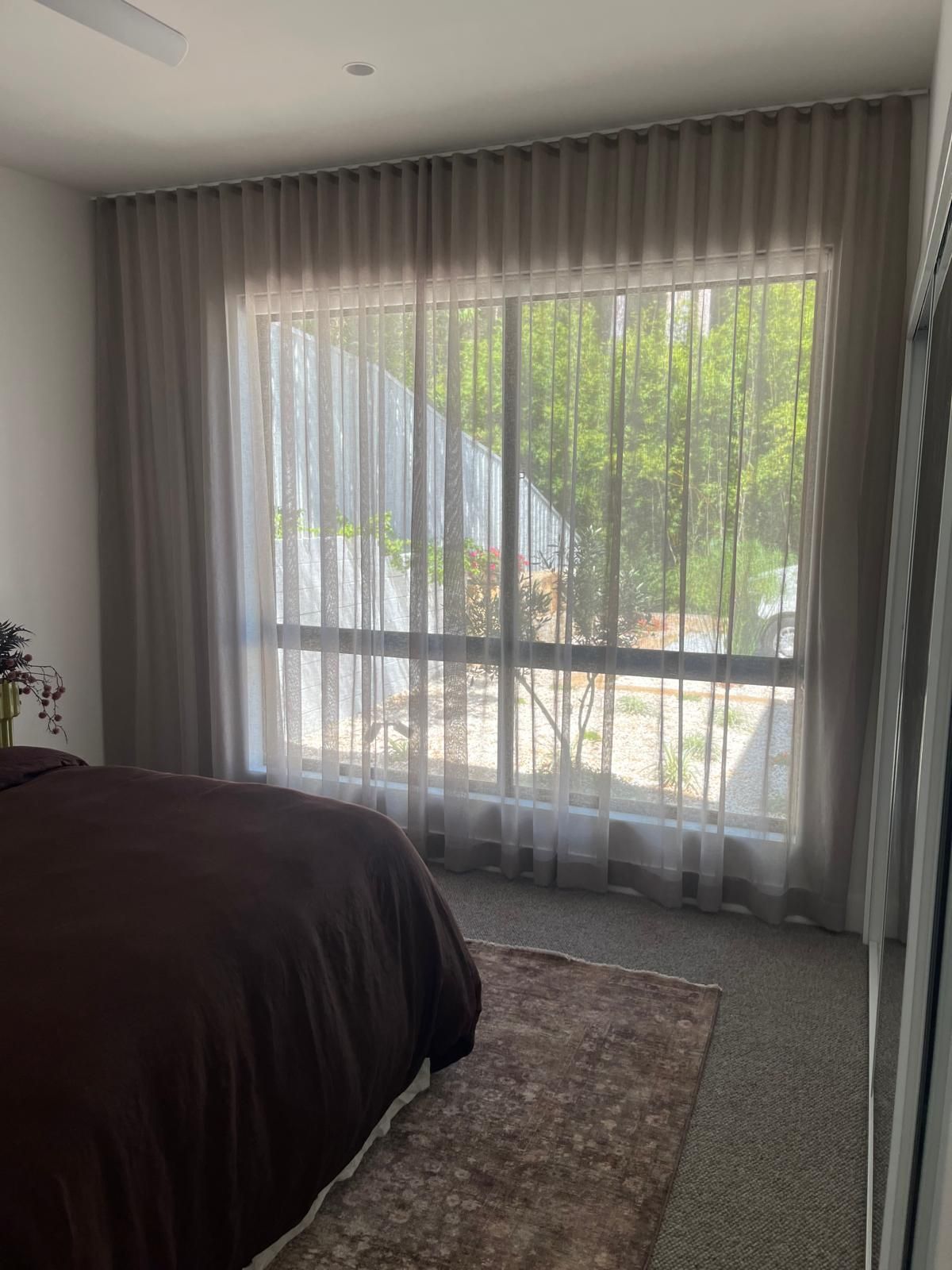 Bedroom with a bed, large window, sheer curtains, and daylight streaming in over a rug.