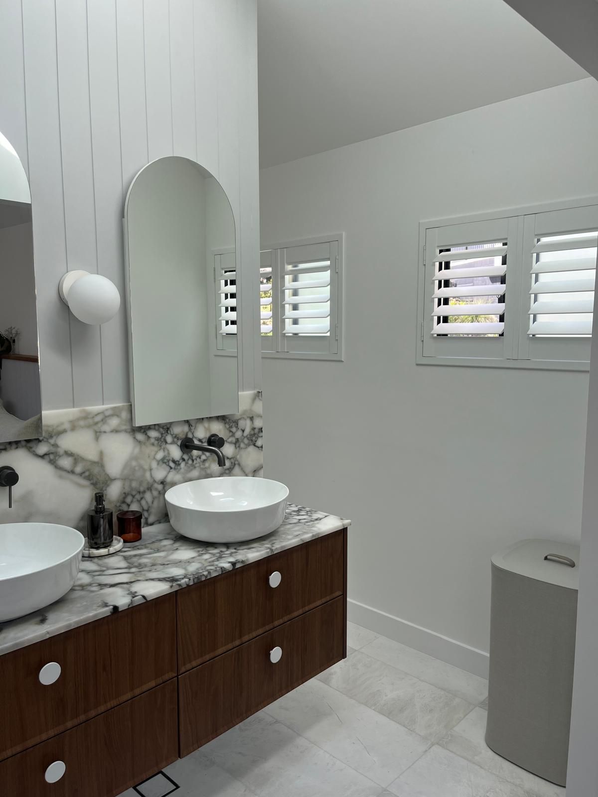 Modern bathroom vanity with marble countertop, vessel sink, wood cabinets, mirror, and white plantation shutters