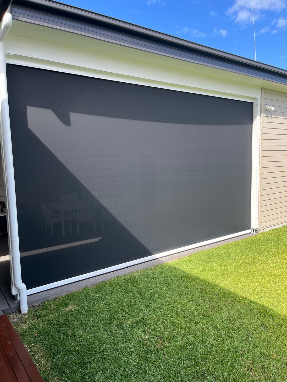 Closed gray outdoor roller shade on a patio opening beside a green lawn under a blue sky